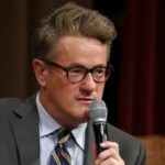 Joe Scarborough speaking at an event, holding a microphone, wearing glasses and a suit
