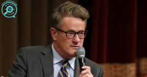 Joe Scarborough speaking at an event, holding a microphone, wearing glasses and a suit