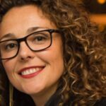 Rishia Haas smiling in a leather jacket and glasses at an evening event, 2025.